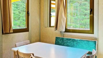 Une pièce avec une table et des chaises, une fenêtre avec des rideaux et une vue sur la verdure à l’extérieur.
