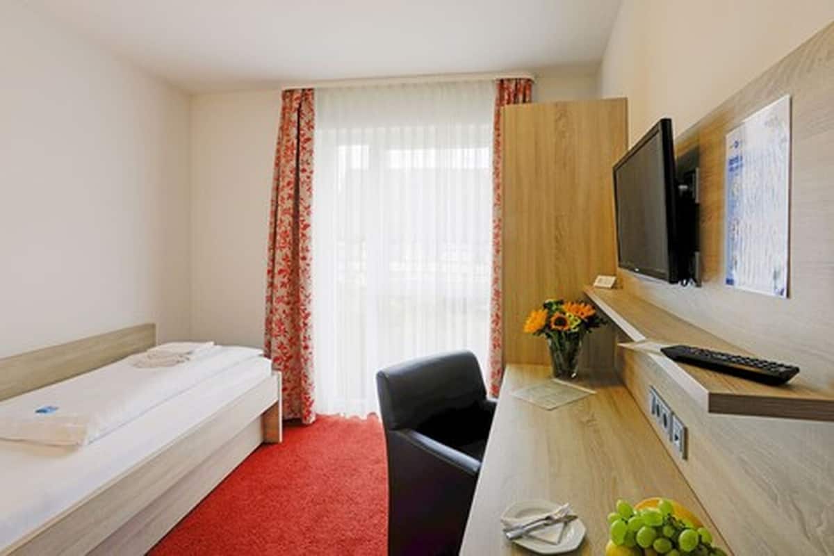 A hotel room with a bed, a desk with a computer, a chair, a TV, and a window with curtains.