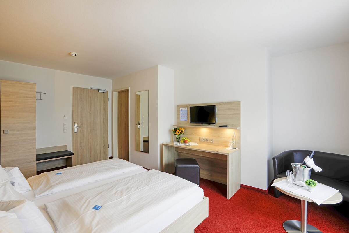 A hotel room with a bed, a desk with a TV, a chair, and a small table with a lamp.