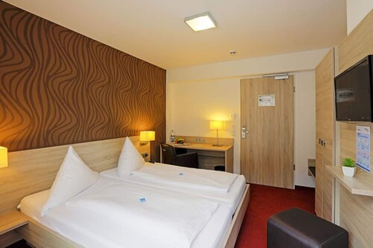 A hotel room with a double bed, a desk, a chair, a TV, and a small plant.