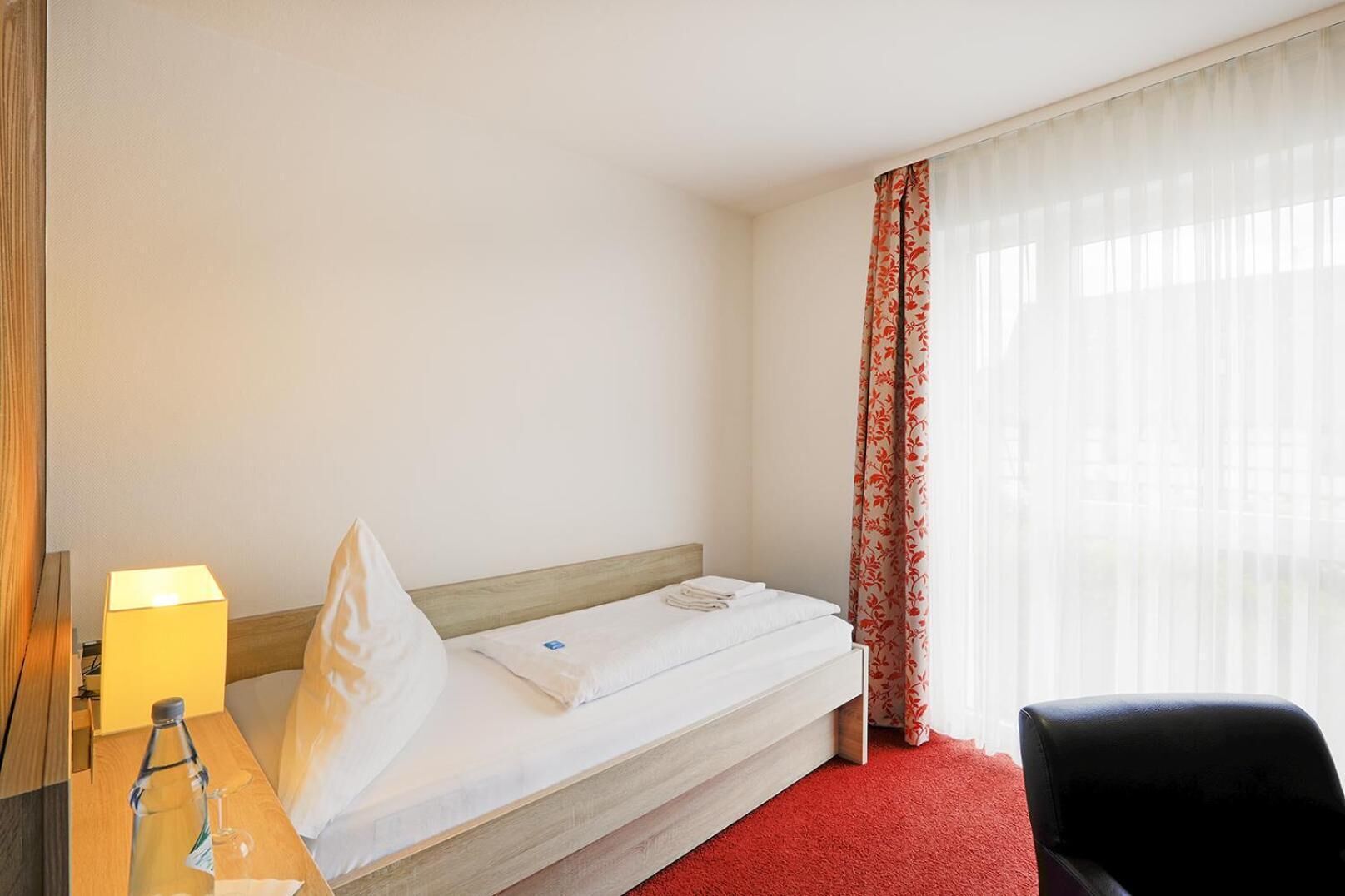Ein Hotelzimmer mit einem Einzelbett, einem Nachttisch mit Lampe, einem Sessel und einem Fenster mit durchsichtigen Vorhängen.