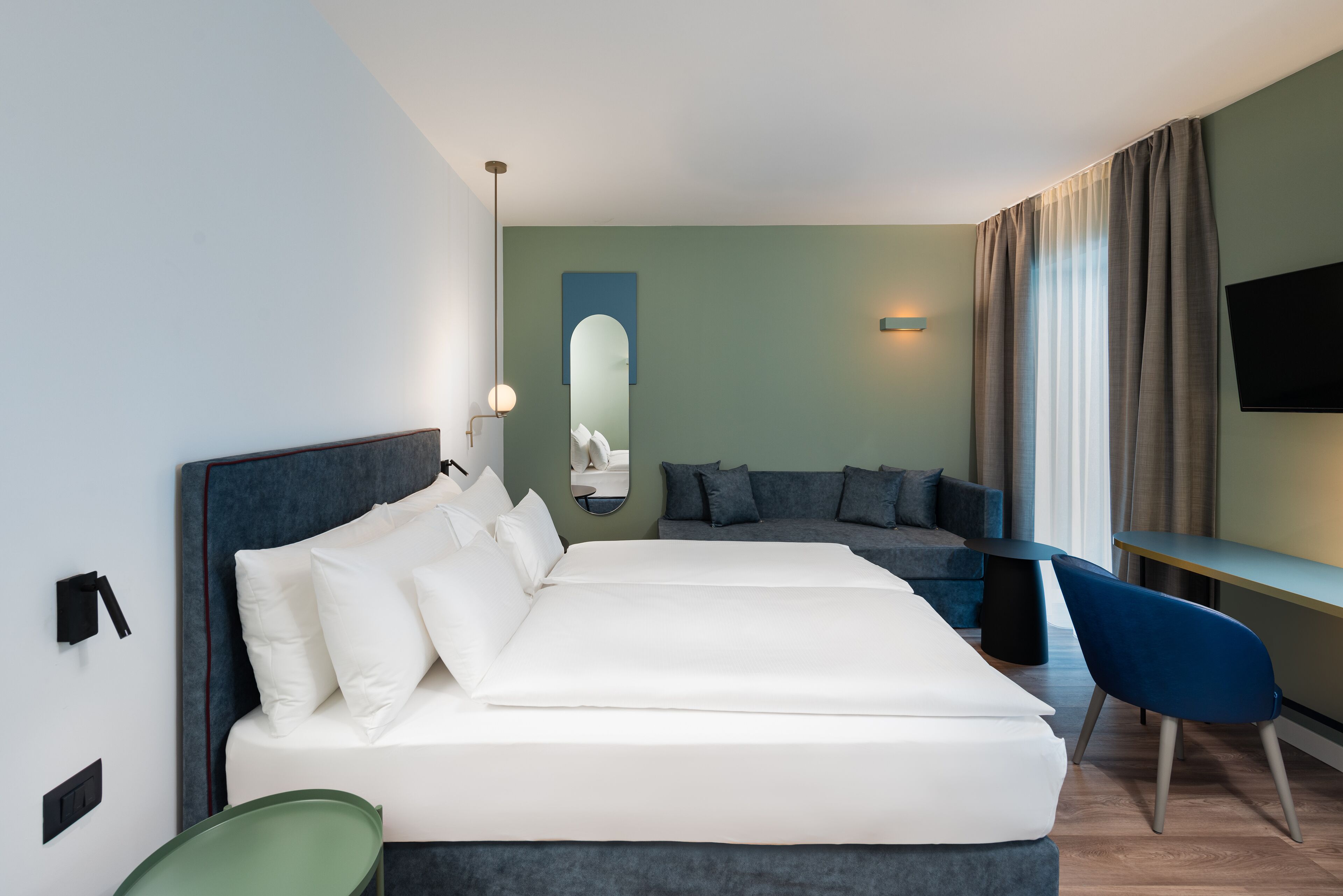 A modern hotel room with a large bed, a seating area with a sofa and chairs, a flat-screen TV mounted on the wall, and a mirror above the bed.