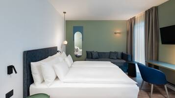 A modern hotel room with a large bed, a seating area with a sofa and chairs, a flat-screen TV mounted on the wall, and a mirror above the bed.