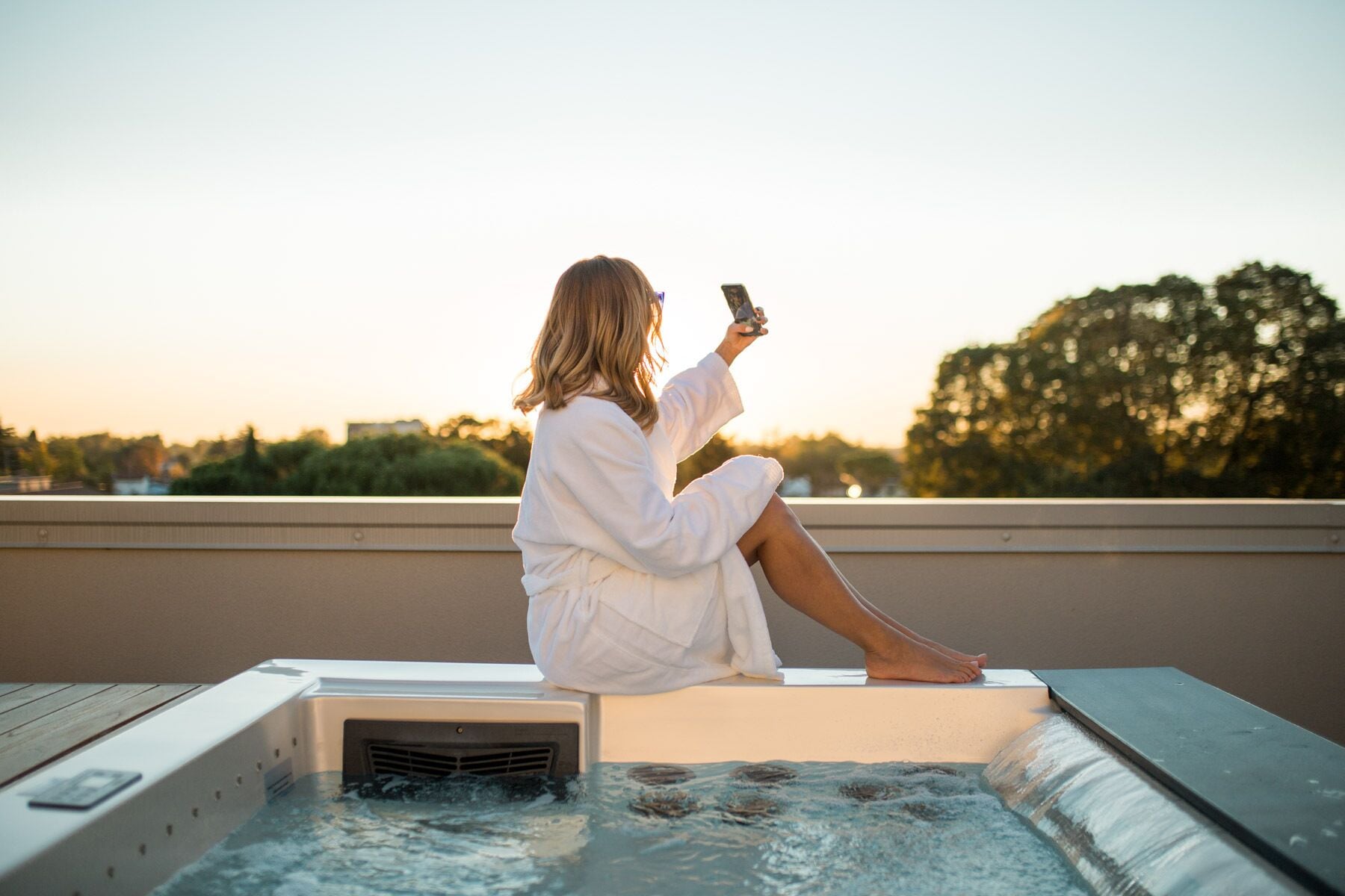 A person sitting on a hot tub with a smartphone, overlooking a cityscape at sunset.