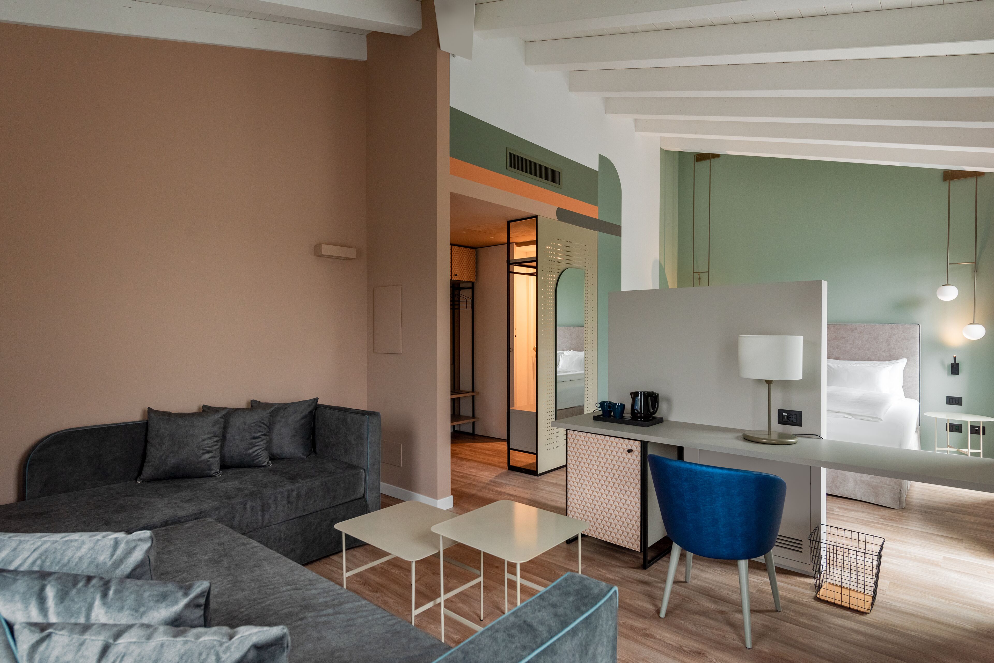 Una habitación de hotel moderna con un sofá gris, una mesita blanca, una silla azul, un escritorio con una lámpara y una cama con ropa de cama blanca.