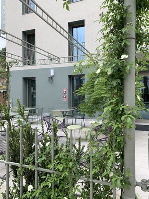 A building with a grey facade, outdoor seating area with green chairs and tables, and a staircase leading to an upper level.