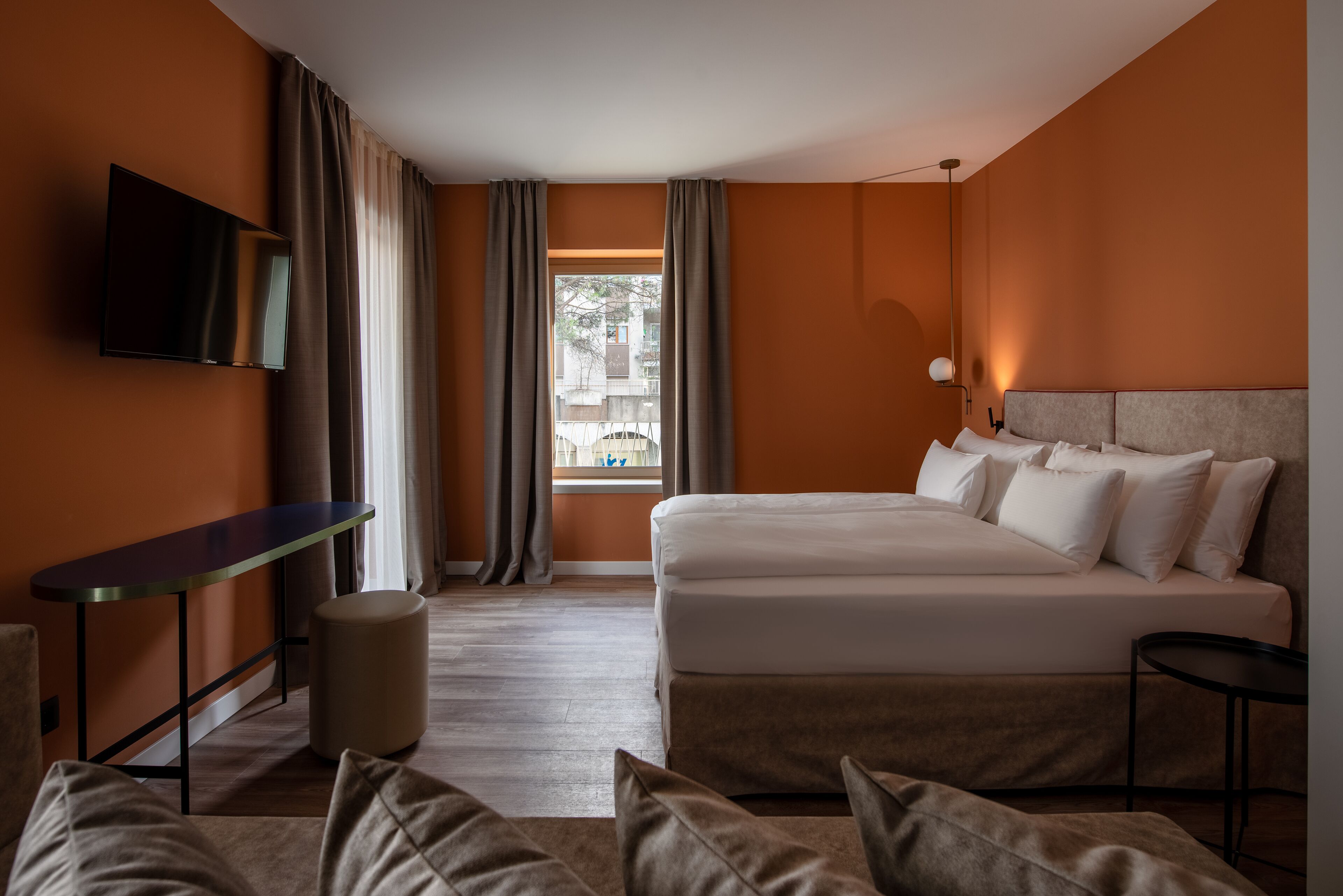 Una camera d'albergo con un letto, una TV a muro, una scrivania e una finestra con tende.
