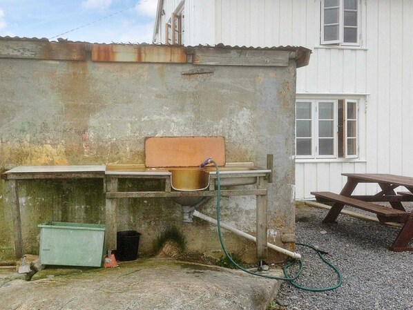 A rustic outdoor kitchen area with a sink, a green trash bin, and a wooden picnic table.