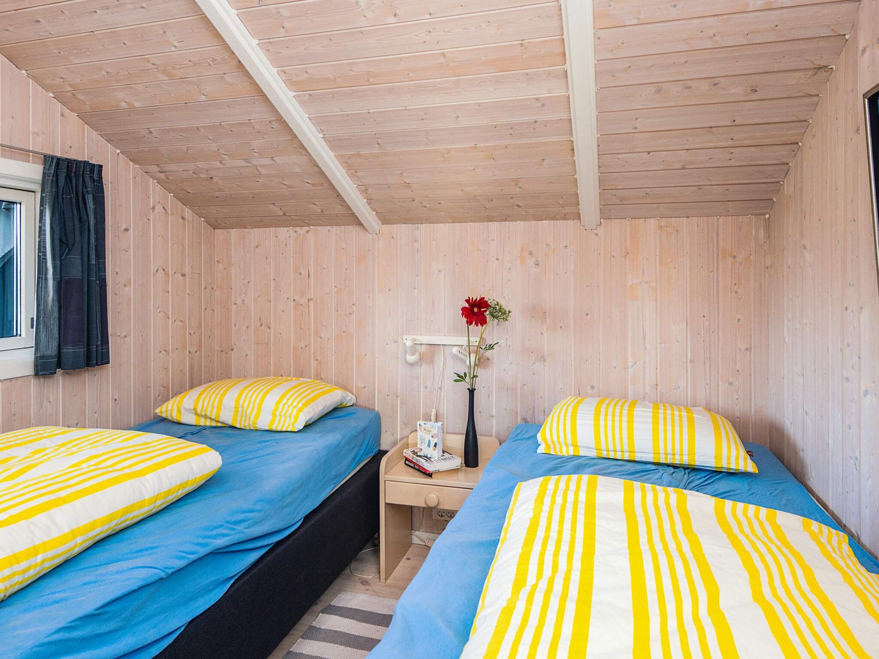A cabin with two beds, each with a blue mattress and yellow-striped pillows, a bedside table with a lamp, and a window with curtains.