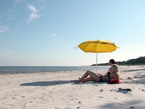 Une personne assise sur une plage de sable, à l’ombre d’un parasol jaune.