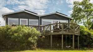 A two-story wooden cabin with a deck and a swing chair, surrounded by greenery. - 7 Person Holiday Home in Knebel-by Traum (Knebel)