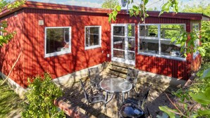 Una cabaña roja de madera con puerta y ventanas de vidrio, un patio con mesa y sillas, y una antena parabólica en el techo.