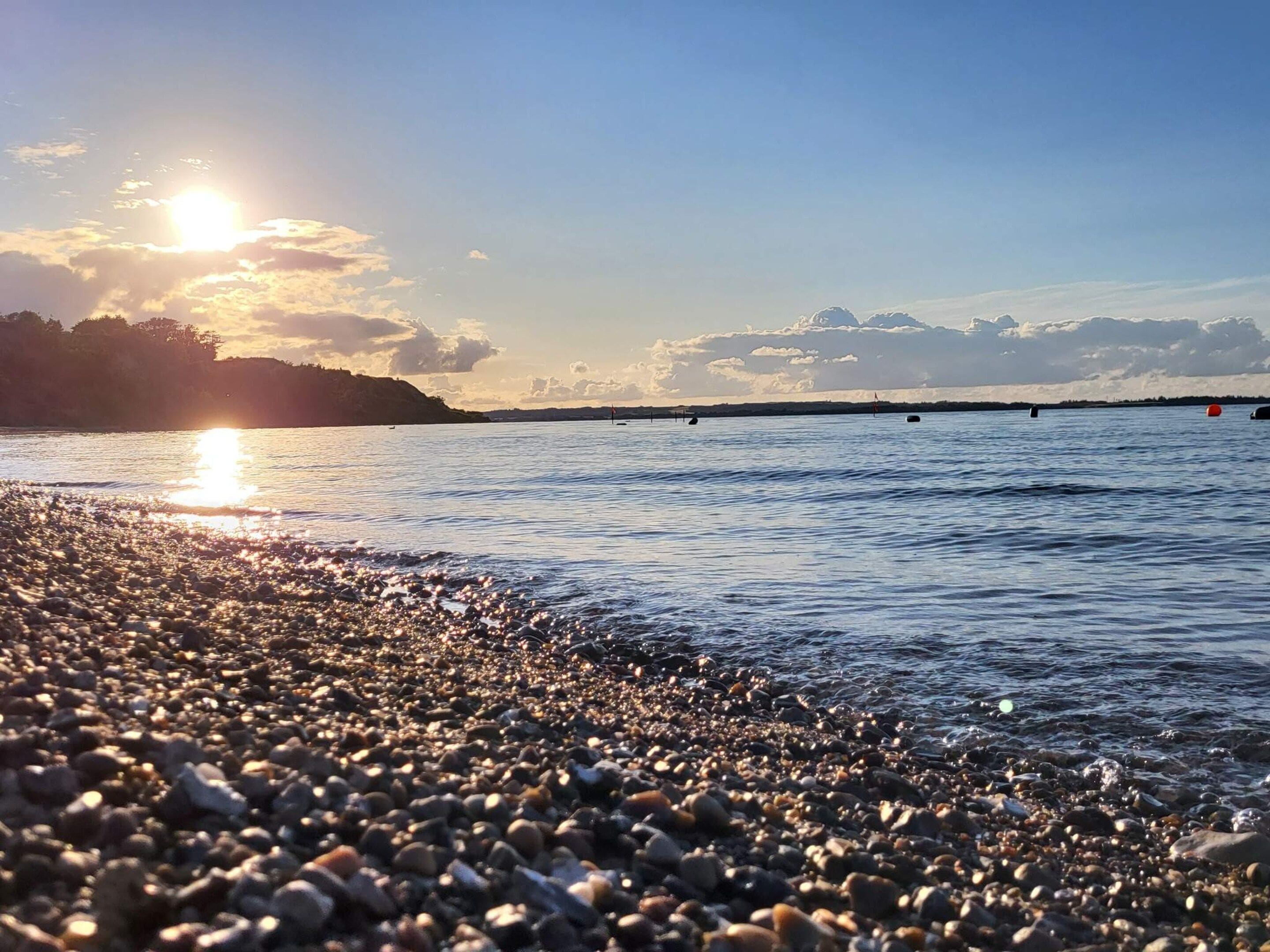 Ein Kiesstrand mit ruhiger See und Sonnenuntergang im Hintergrund.