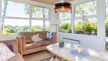 Una habitación acogedora con techo de madera, un sofá, una mesa con lámpara y ventanas amplias que dan a zonas verdes.