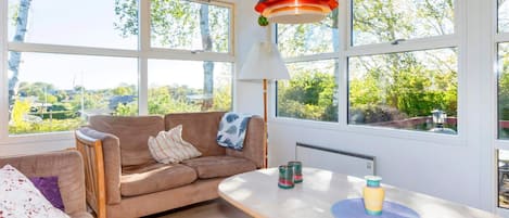 Una habitación acogedora con techo de madera, un sofá, una mesa con lámpara y ventanas amplias que dan a zonas verdes.