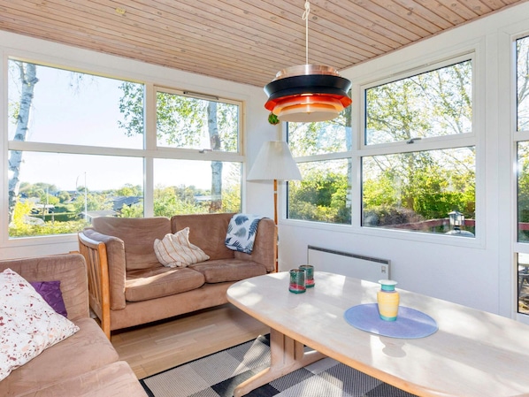 Quarto aconchegante com teto de madeira, um sofá, uma mesa com abajur e janelas amplas com vista para o verde.