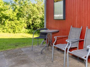 Un área de estar al aire libre con sillas de metal y una mesa, un edificio rojo y una zona verde con árboles.
