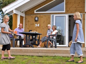 A wooden house with a number '54' on the wall, a patio with a table and chairs, and a person playing with a ball.