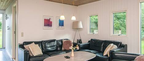 Un salón moderno con un sofá de cuero negro, una mesa redonda de centro y una lámpara de pie. Hay dos ventanas que ofrecen luz natural y vistas a espacios verdes.