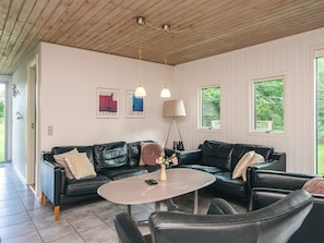 A modern living room with a black leather sofa, a round coffee table, and a floor lamp. There are two windows providing natural light and a view of greenery outside. - 8 Person Holiday Home in Orsted (Ørsted)