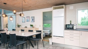 A modern kitchen with a dining area, featuring a wooden ceiling, a dining table with chairs, a built-in oven, and a window with a view of greenery.