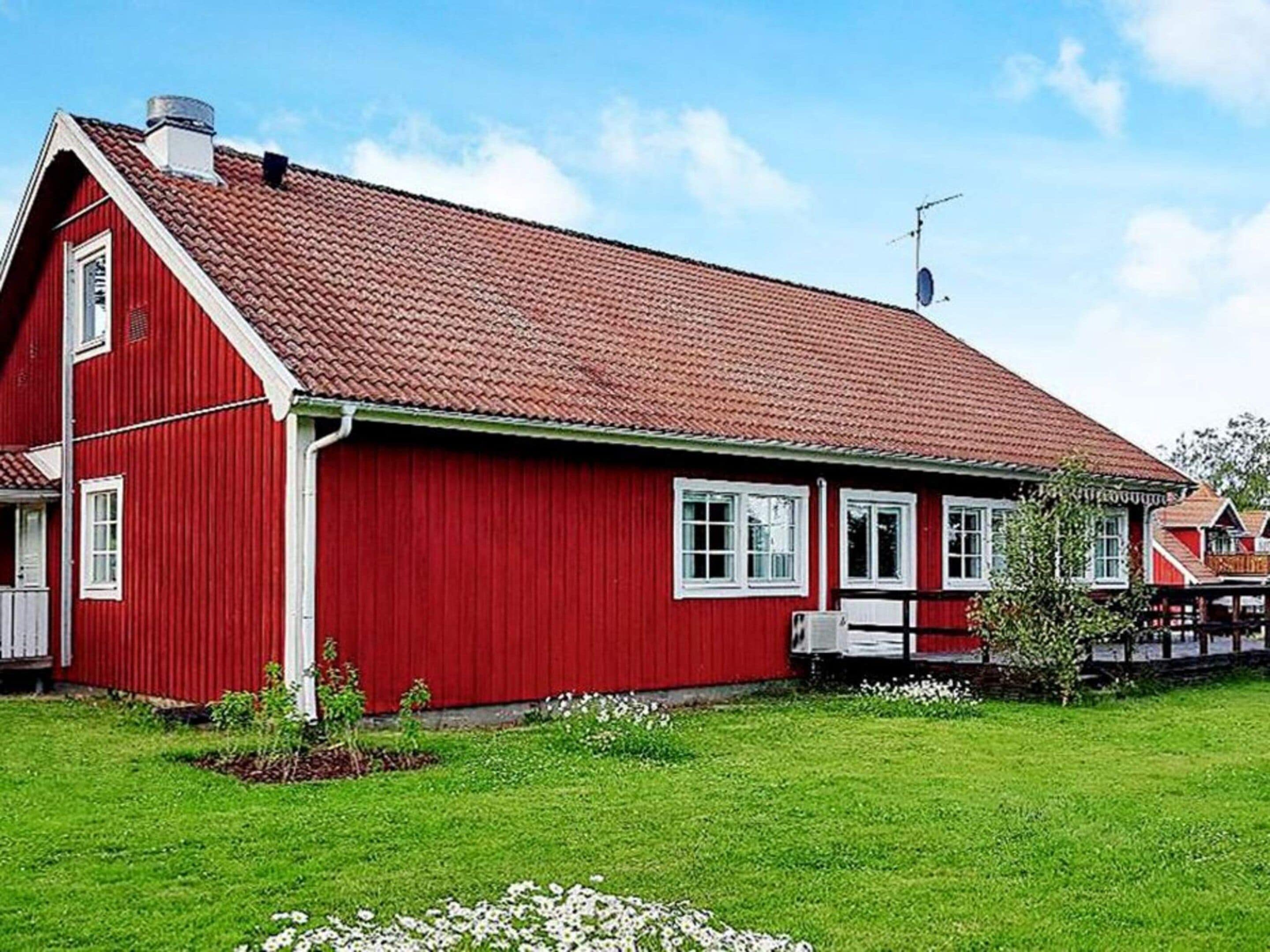 A red wooden house with a sloped roof, white windows, and a small garden with white flowers.