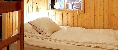 A bunk bed with a single mattress and a pillow, wooden walls, a window with a curtain, and a lamp.