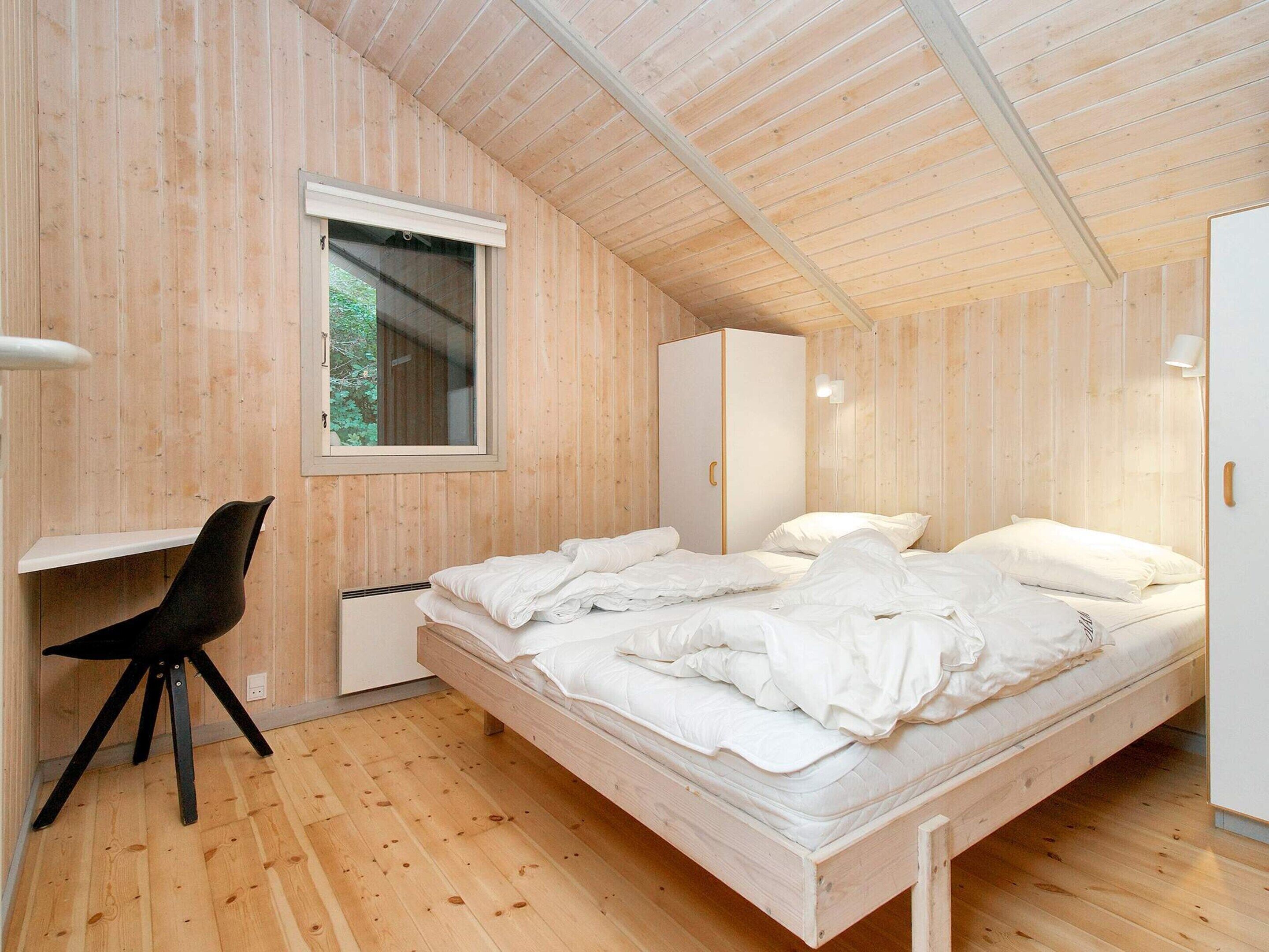 Quarto com piso de madeira, cama com roupa de cama branca, escrivaninha com cadeira e guarda-roupa.
