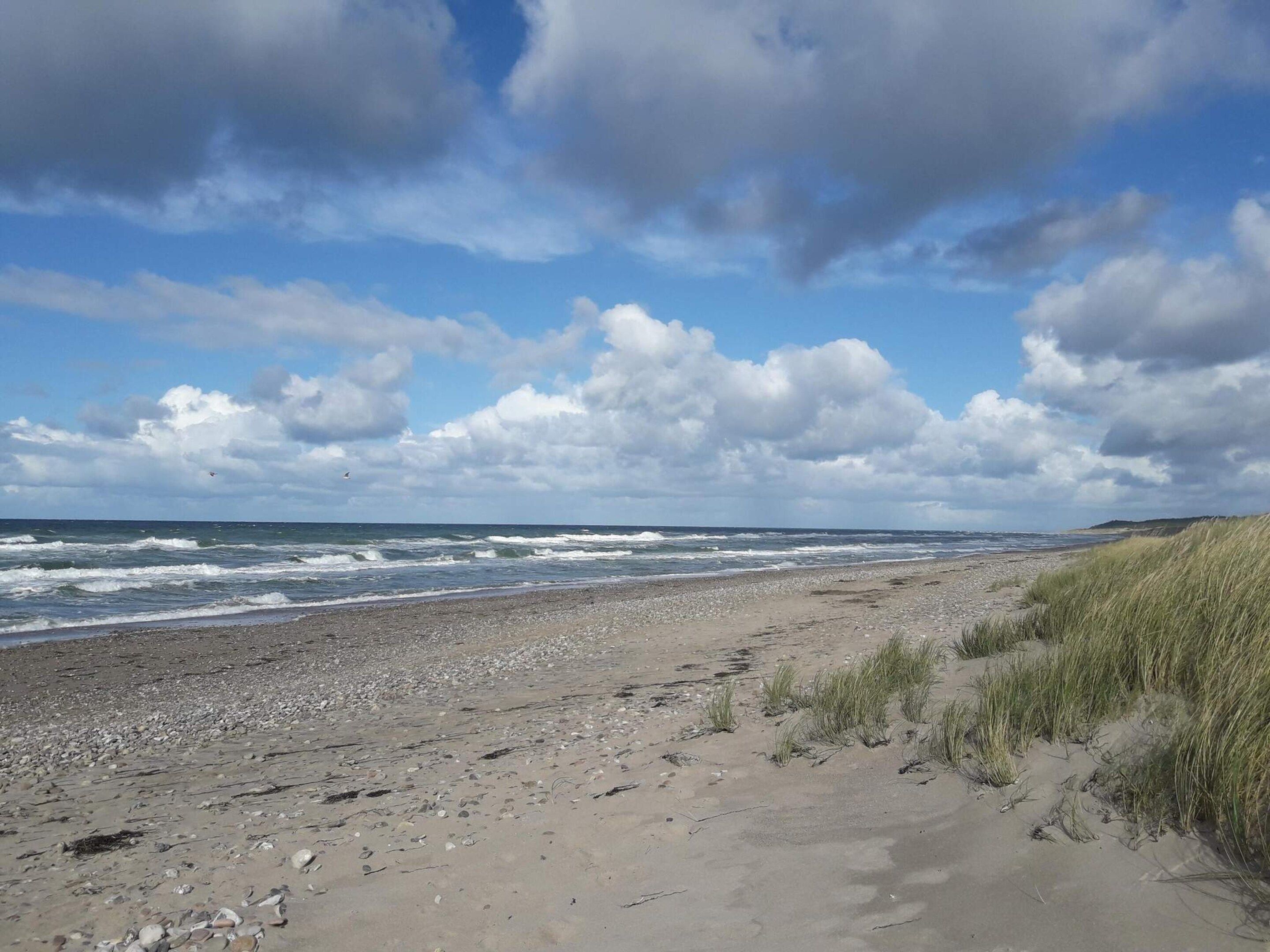 Una playa con orilla pedregosa, dunas cubiertas de hierba y un cielo parcialmente nublado.