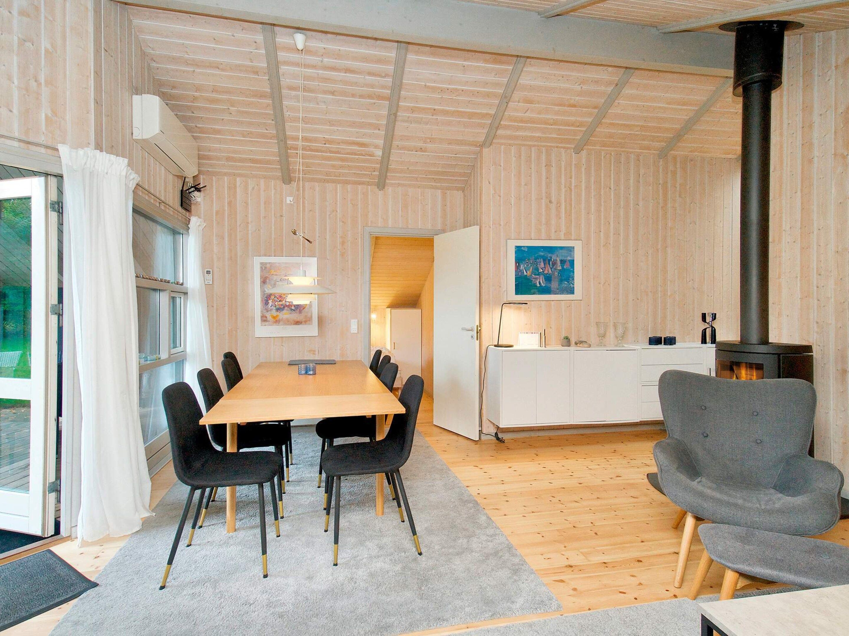 A modern interior with a wooden floor, a dining table with chairs, a gray armchair, a white cabinet, and a black stove.