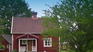 Et rødt træhus med tegltag, en veranda med hvide gelændere og en lille græsplæne med et bord og stole.