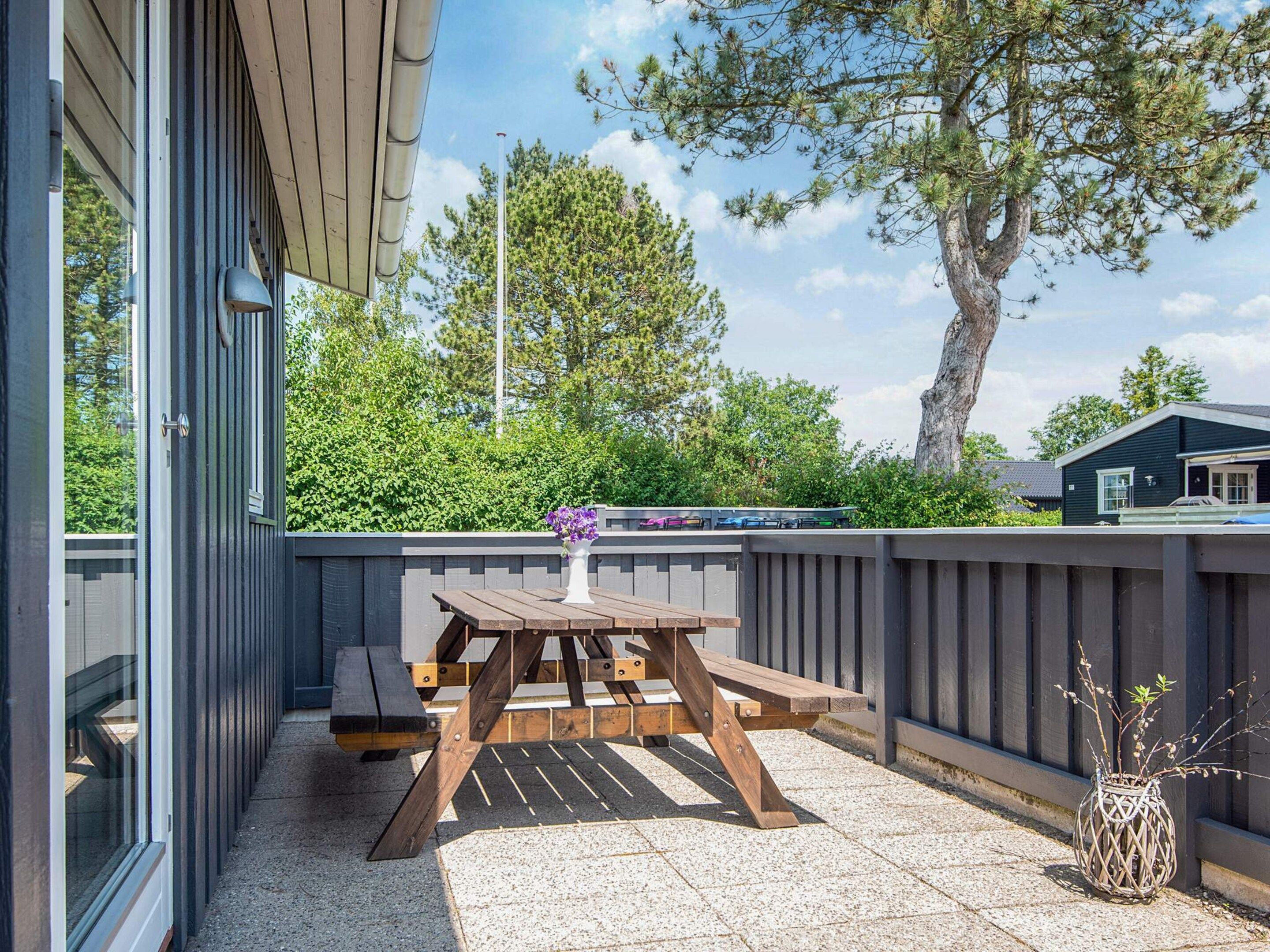 A patio with a wooden table and benches, a small potted plant, and a view of trees and a building in the background.