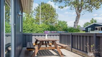 A patio with a wooden table and benches, a small potted plant, and a view of trees and a building in the background.