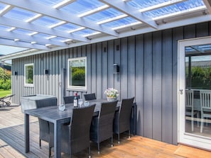 A covered patio with a table and chairs, a glass door, and a view of greenery.