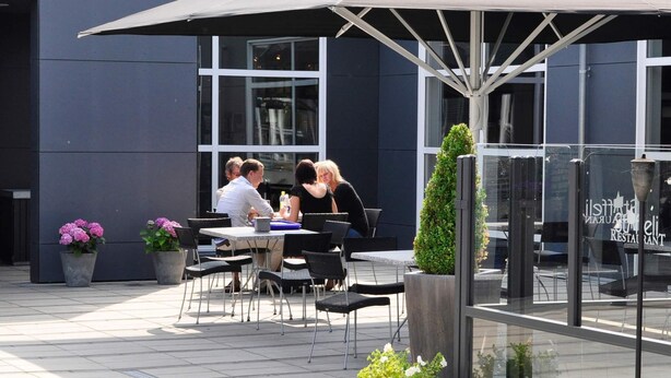 Un café al aire libre con mesas y sillas bajo un paraguas grande, rodeado de plantas en macetas y un edificio moderno al fondo.