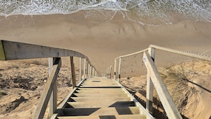 En trætrap fører ned til en strand med sand og bølger i baggrunden.