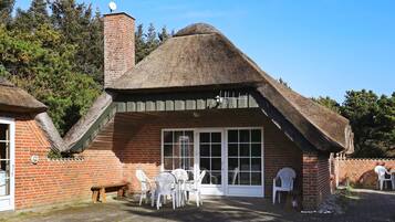 A thatched-roof cottage with a brick exterior, a large window, and a patio with white plastic chairs.