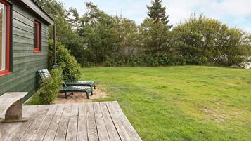 A wooden deck with a bench and a table, a grassy yard with trees, and a building with green siding and red trim.
