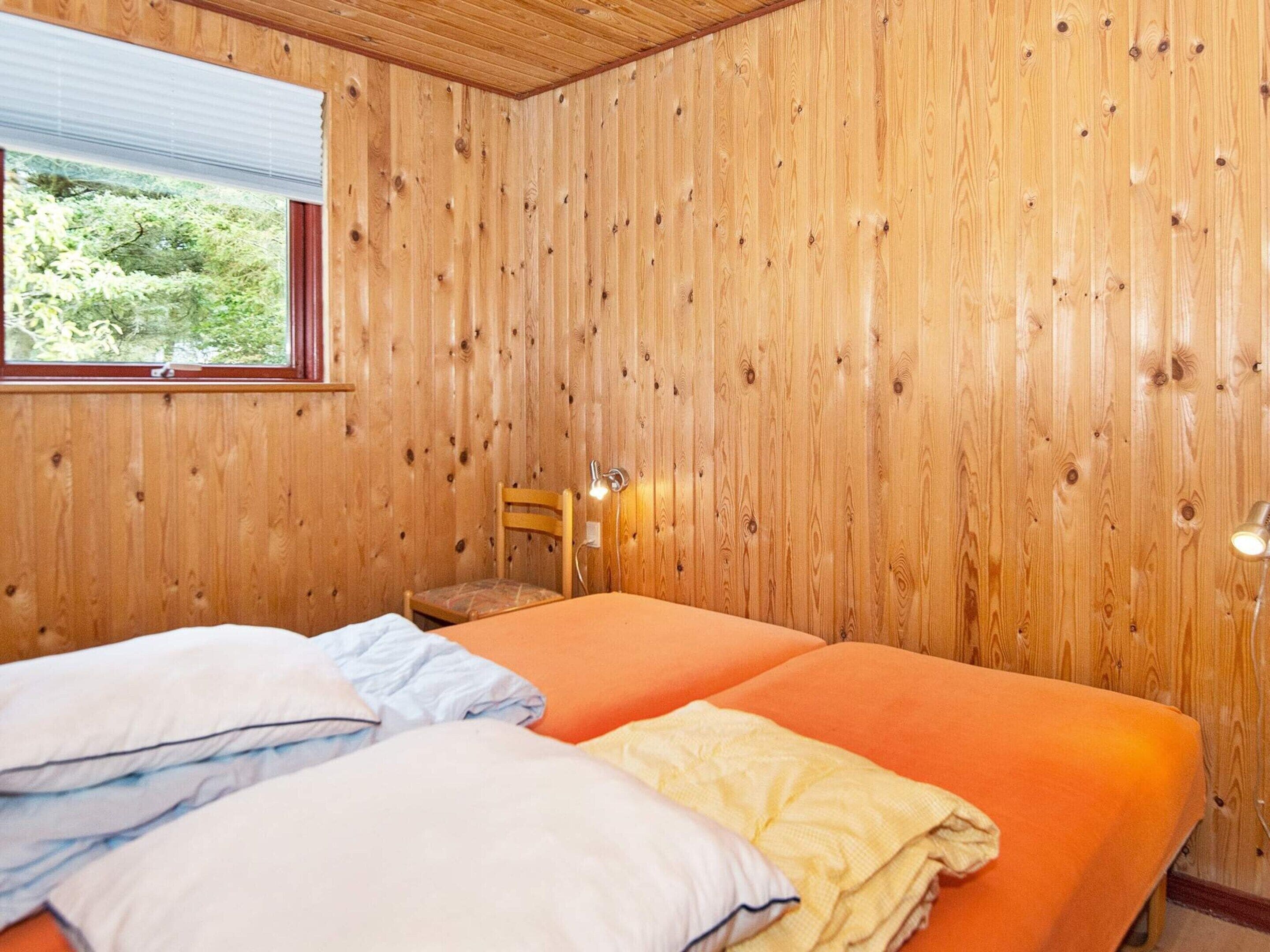 A room with wooden panel walls, a bed with orange bedding, a window with a view of greenery, and a chair with a lamp.