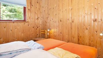 A room with wooden panel walls, a bed with orange bedding, a window with a view of greenery, and a chair with a lamp.