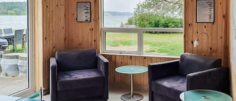 Une pièce aux murs en bois, avec deux fauteuils sombres, une table de ping-pong et une vue sur un lac et la verdure environnante.