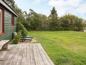 A wooden deck with a bench and a table, a grassy yard with trees, and a building with green siding and red trim. - Spacious Holiday Home in Storvorde With Sea Nearby (Skanderborg)