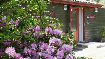 Un edificio con puerta roja y ventanas, rodeado de follaje verde y flores rosadas.