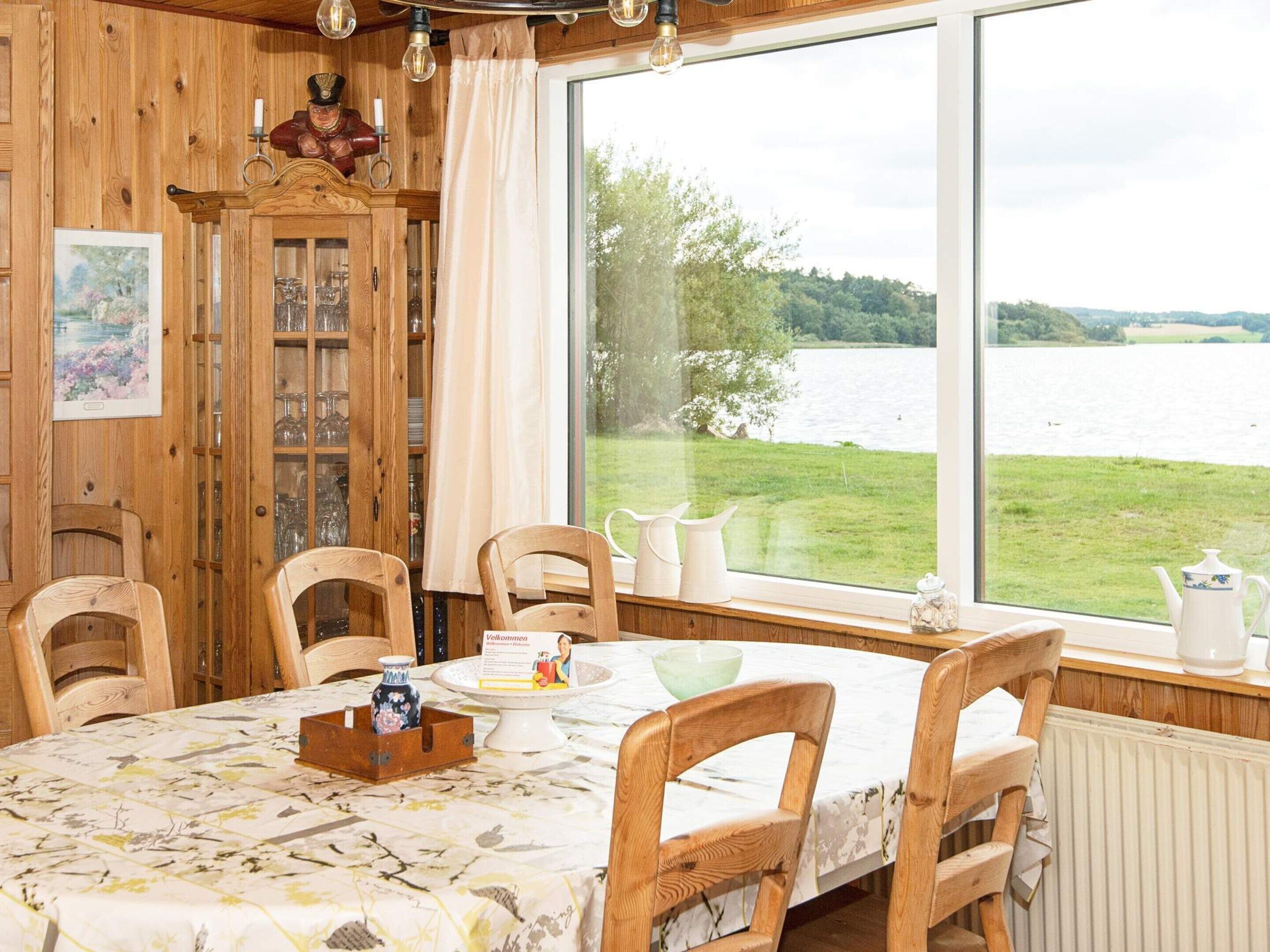A dining area with a table set for four, wooden chairs, a cabinet with glass doors, and a view of a lake through large windows.