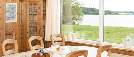 Et spiseområde med et bord dækket til fire personer, træstole, et skab med glasdøre og udsigt til en sø gennem store vinduer.