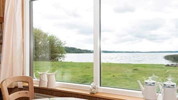 A room with a large window overlooking a grassy area and a body of water. There is a table with a bowl and teapots on the windowsill.