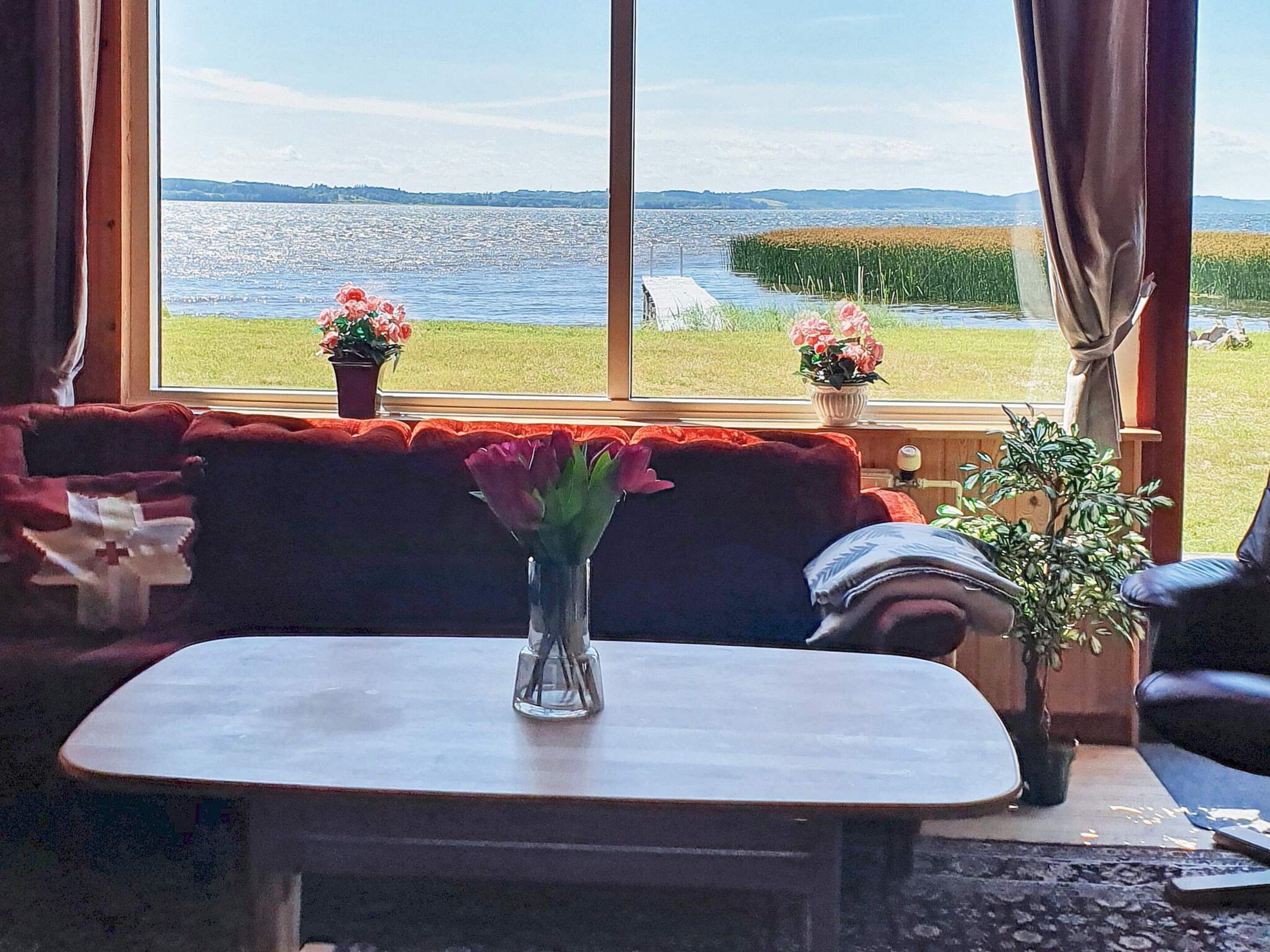 A room with a view of a body of water, a red sofa, a small table with a vase of flowers, and a potted plant.