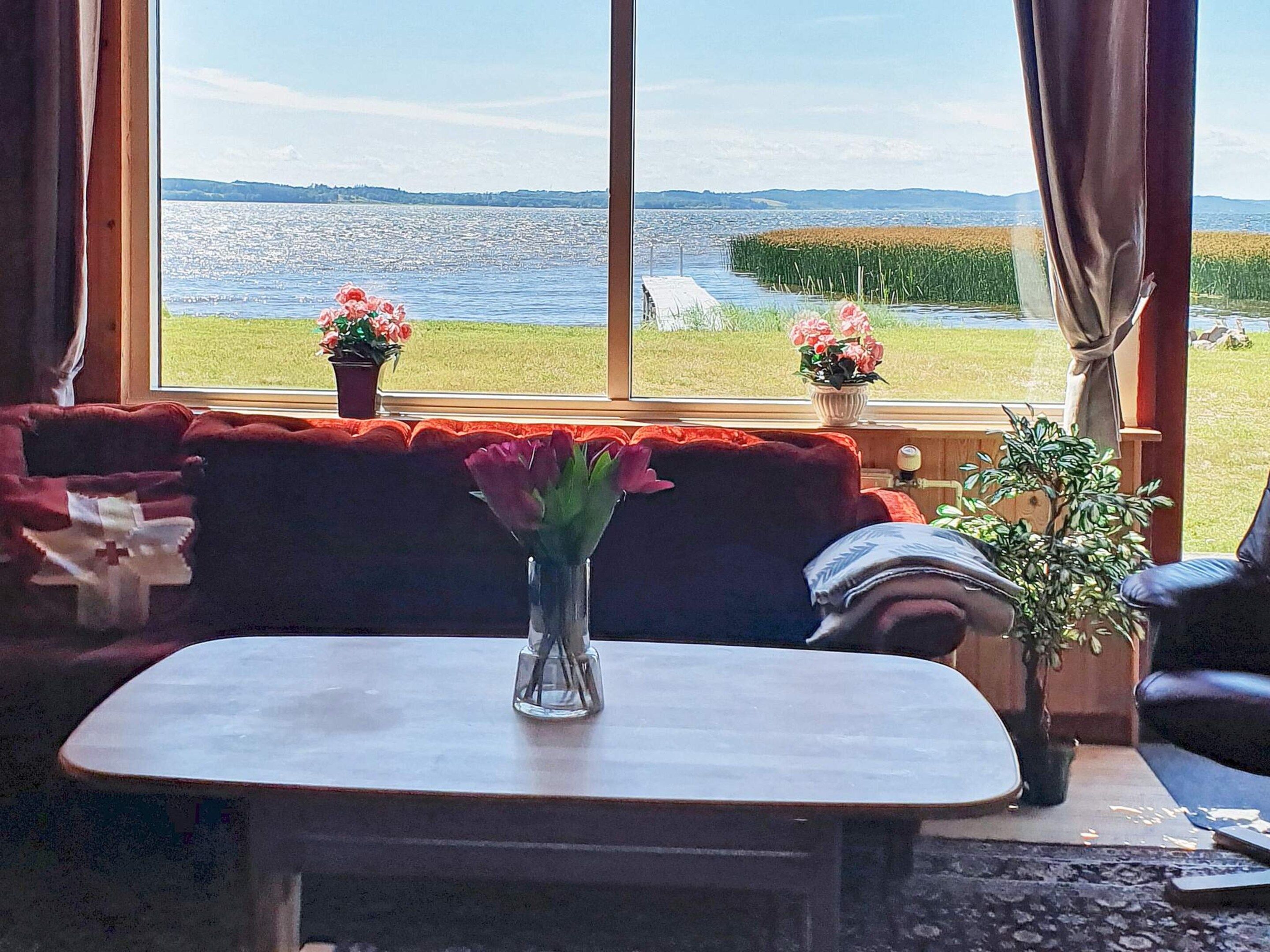 A room with a large window, a red sofa, a small table with a vase of flowers, and a potted plant.