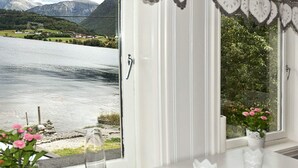 Ein Zimmer mit einem großen Fenster, das Blick auf einen See und Berge bietet. Auf der Fensterbank stehen zwei Blumentöpfe mit rosa Blüten.