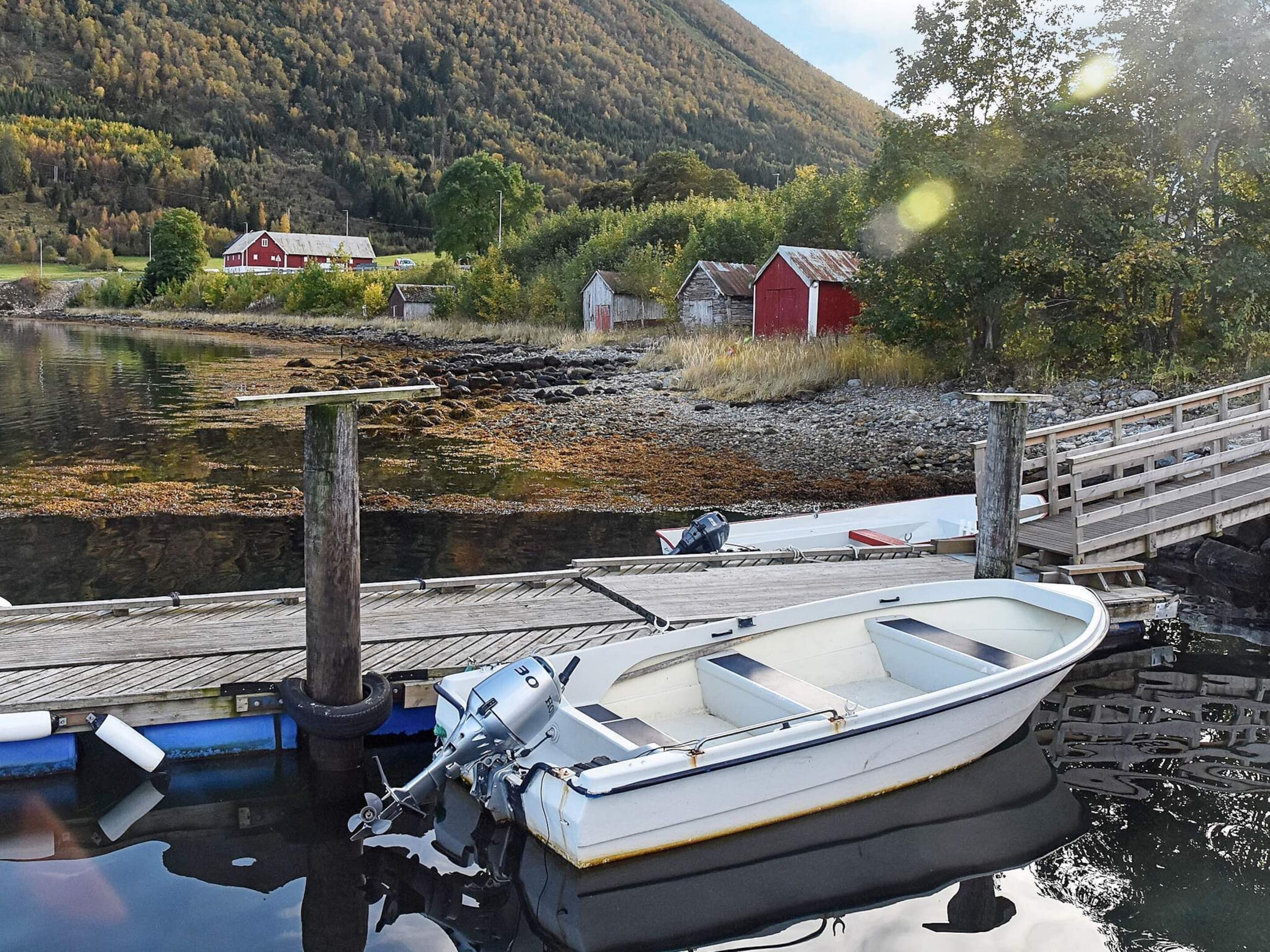 A small boat docked at a wooden pier with a rocky shoreline and a red building in the background.